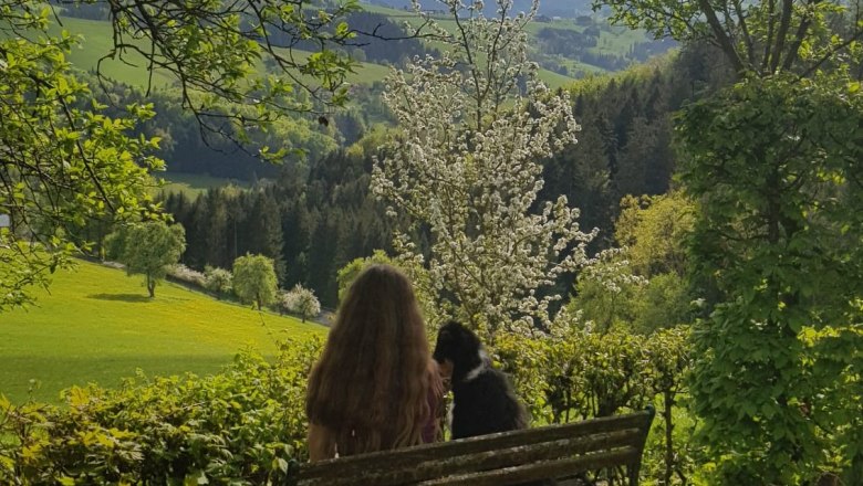 Aussicht mit Hund, © Andrea Stiftner A woman is sitting on a bench with a dog, looking out at a green landscape with blossoming trees and mountains in the background.