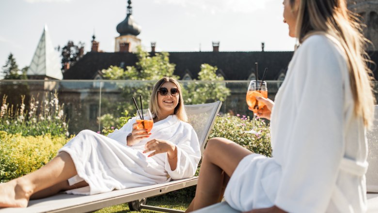 The castle on the Iron Road, © Dominik Stixenberger Two women in bathrobes relax on deckchairs with drinks in front of a castle.