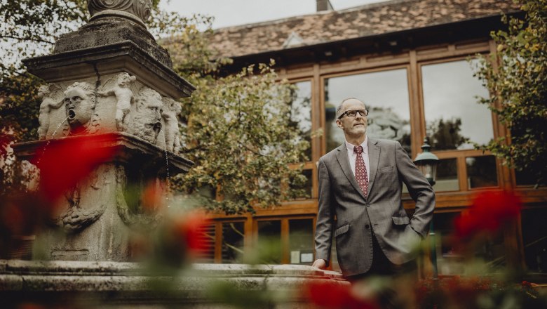 Andreas Horvath, © Niederösterreich Werbung/Sophie Menegaldo A man in a suit stands next to a stone fountain with gargoyles, surrounded by red flowers and a building in the background.