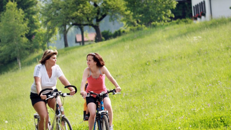 St. Aegyde in Neuwalde, © Mostviertel Tourismus/Weinfranz.at Two women ride bicycles through a green landscape with hills and trees in the background.