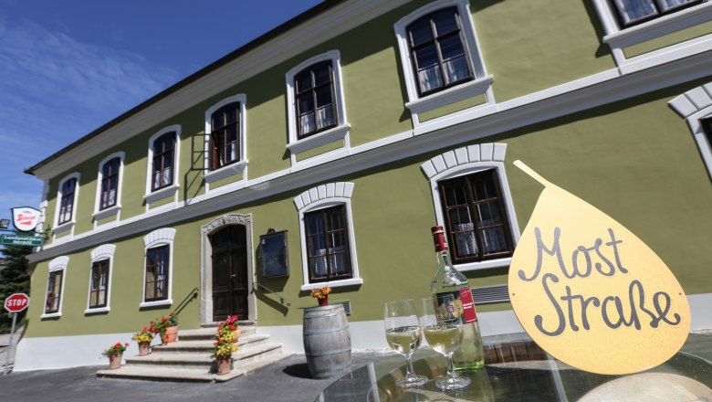 Berndl's Landgasthaus, © schwarz-koenig.at Green building with white windows, in front of it a table with a bottle of wine and glasses, sign with the inscription 'Cider Straße'.