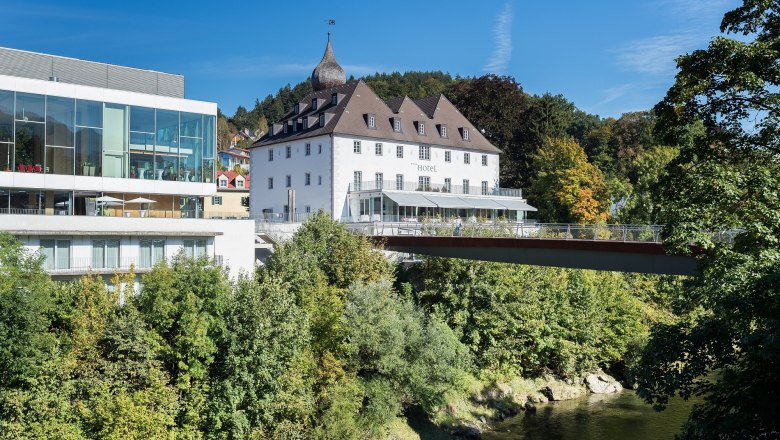 Castle on the Iron Road, © Stixenberger A modern building next to a historic castle, surrounded by trees and a river, under a blue sky.