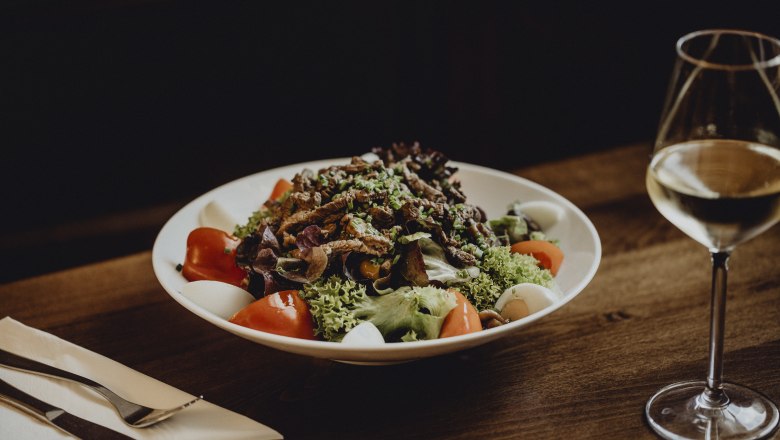 Heiligenkreuz specialty salad, © Niederösterreich Werbung/Sophie Menegaldo A plate of mixed salad, tomatoes, eggs and strips of meat on a wooden table, with a glass of white wine next to it.