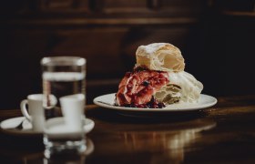 Monastery cream slice, © Niederösterreich Werbung/Sophie Menegaldo A plate with a monastery cream slice, garnished with red berries, stands on a wooden table. A glass of water and two espresso cups can be seen in the background.