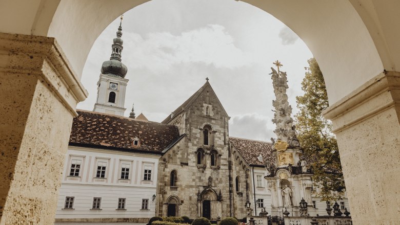 Inn, monastery and monastery store, © Niederösterreich Werbung/Sophie Menegaldo View through an archway to a historic monastery building with a tower and a baroque column in the foreground.