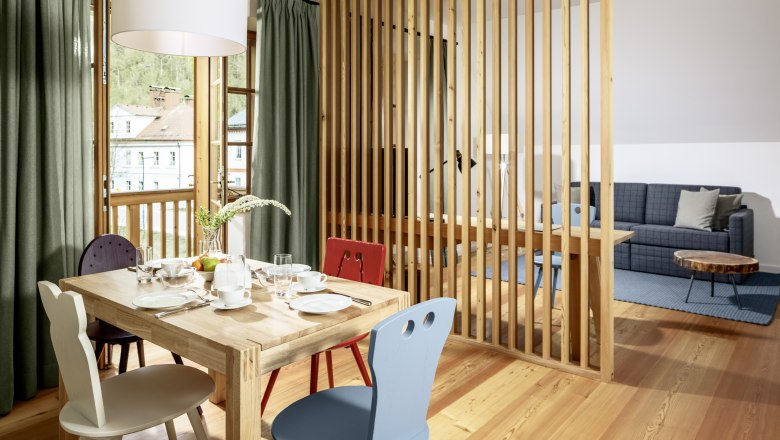 Hotel zum Glockenturm, © www.croce.at A bright hotel room with wooden floor, dining table and chairs, separated by a wooden slatted wall, with a view of a sofa and a coffee table.