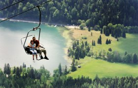 Gemeindealpe Mitterbach, © weinfranz.at Two people sit in a chair lift above a green landscape with a lake in the background.