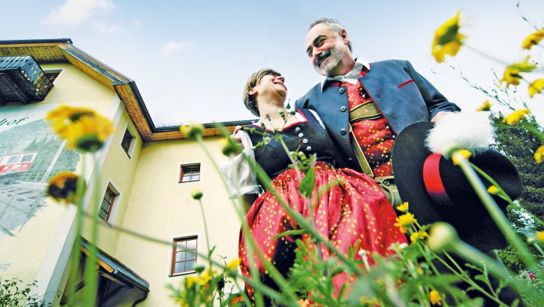 Steinschalerhof Nature Hotel, © weinfranz.at A couple in traditional costume stand in front of a yellow building surrounded by flowers, smiling at each other.