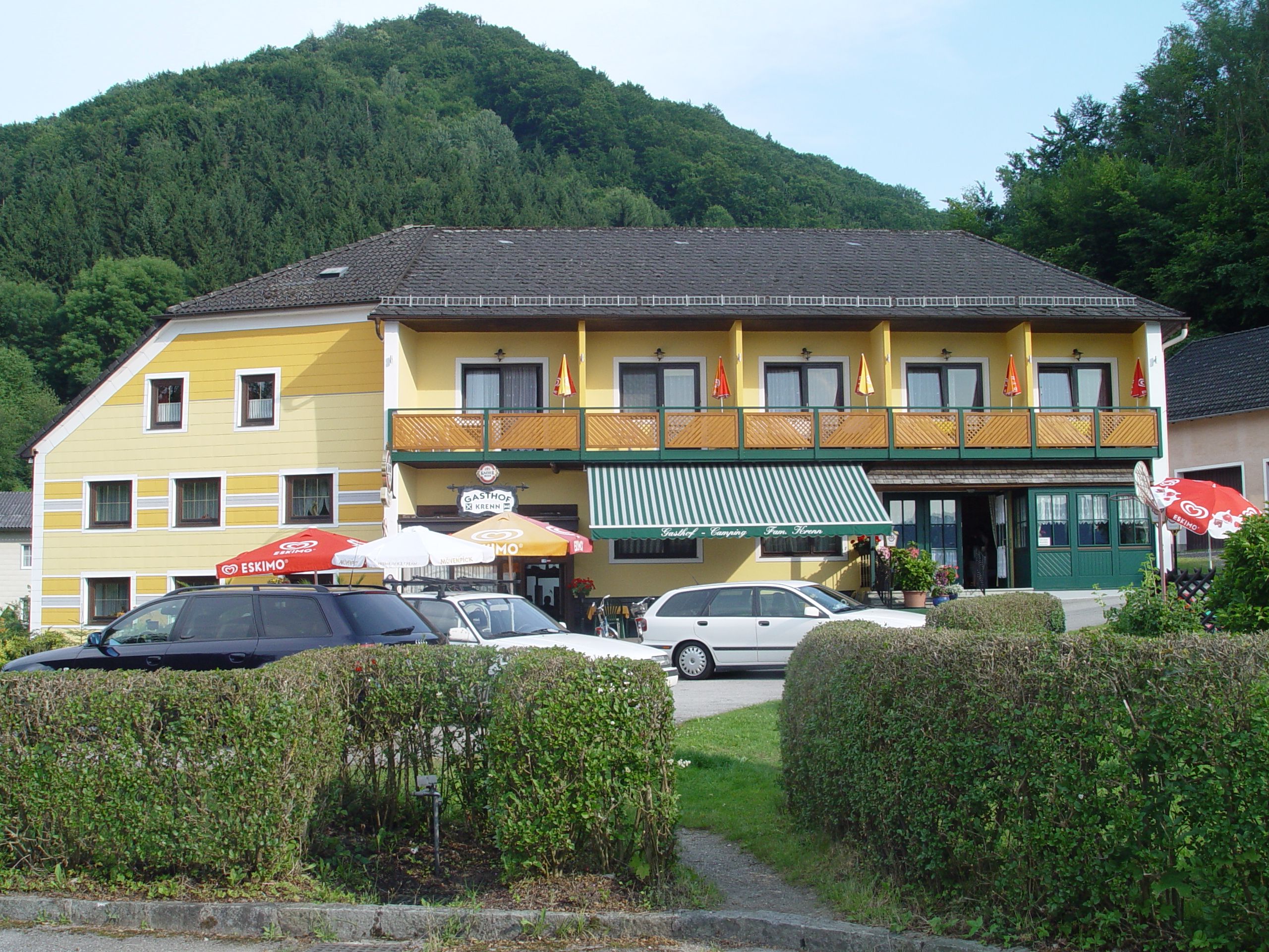 Yellow guesthouse with balcony and parasols in front of a wooded hill.