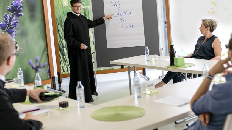 A seminar at Seitenstetten Abbey with a lecturer dressed as a monk pointing at a flipchart. Participants sit at tables.