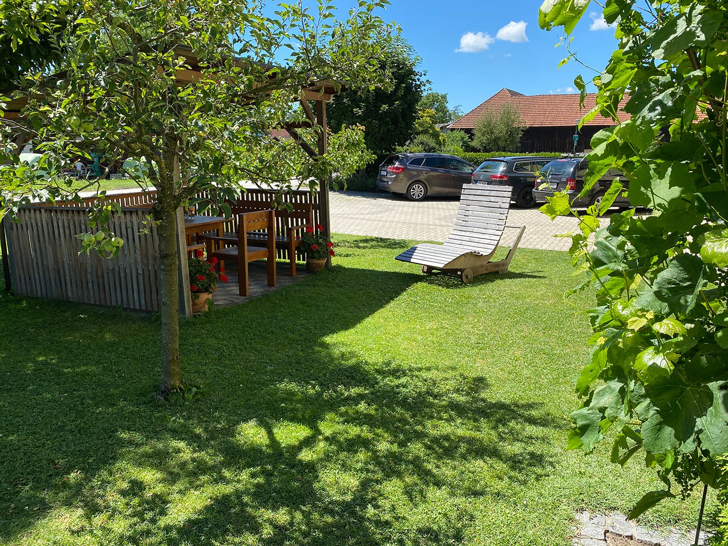 Garden with pergola, wooden table and chairs, sun lounger on lawn, cars in the background.
