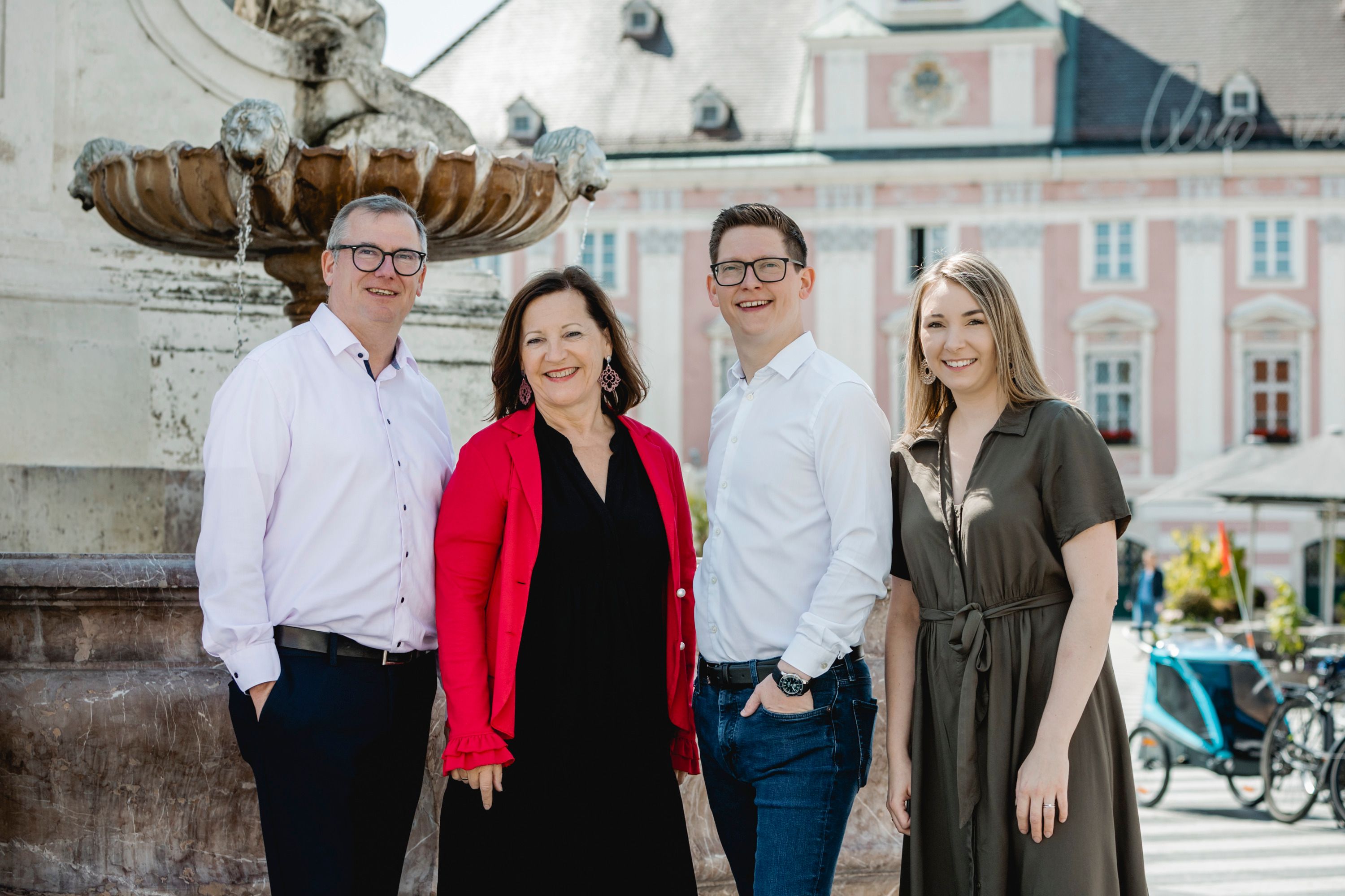 Four people stand smiling in front of a fountain in an urban setting.