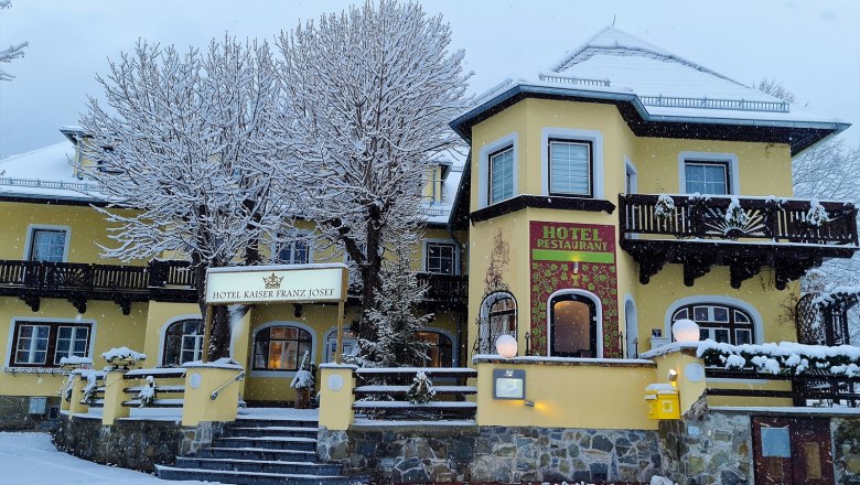 A yellow hotel with the name Kaiser Franz Josef in winter, surrounded by snow-covered trees and a snowy forecourt.