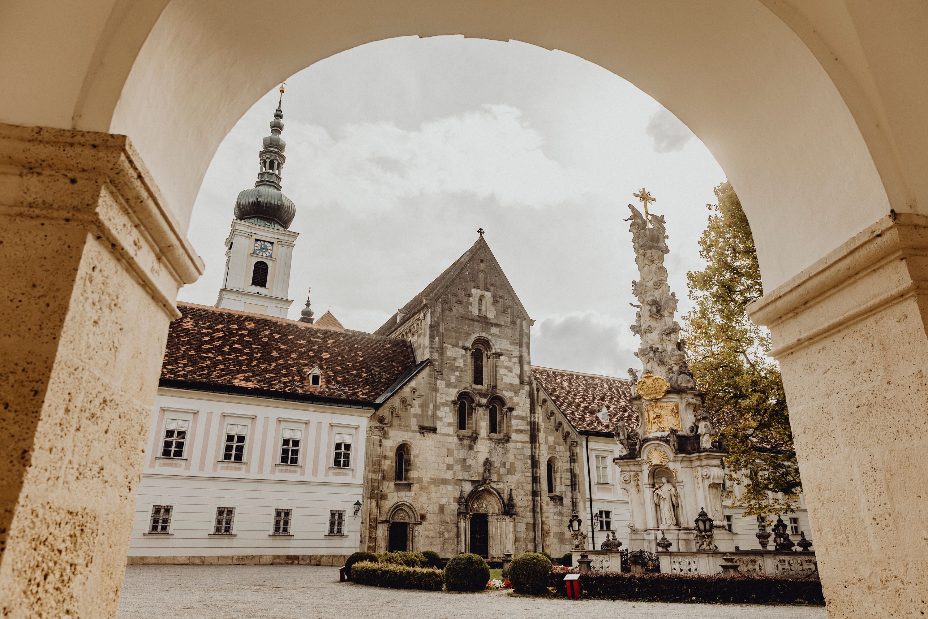 View through an archway to a historic monastery building with a tower and a baroque column in the foreground.