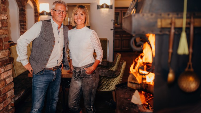 A man and a woman stand smiling in a cozy room with a fireplace.