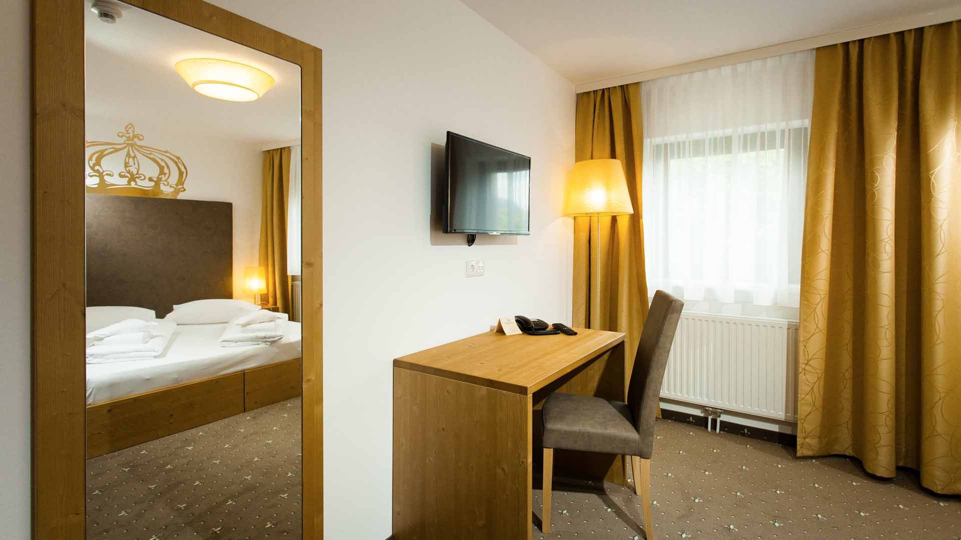 A hotel room with a double bed, a desk and chair, a TV on the wall and yellow curtains.