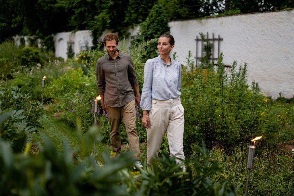 A man and a woman walk through a green garden surrounded by plants.