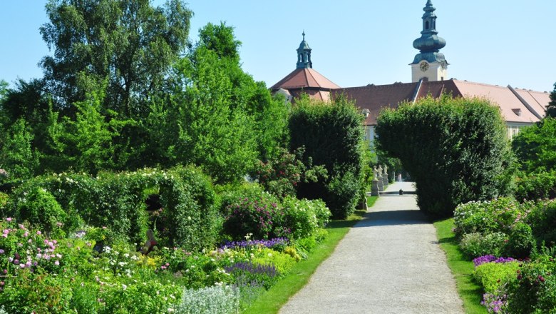 A well-kept garden path with flowers and trees, in the background a historic building with towers.
