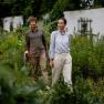 A man and a woman walk through a green garden surrounded by plants.