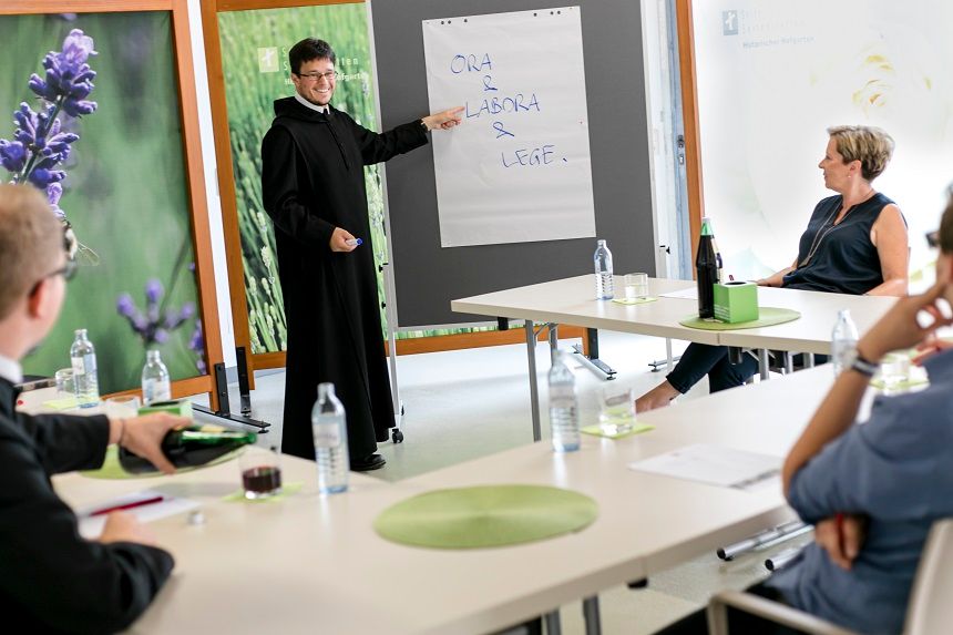 A seminar at Seitenstetten Abbey with a lecturer dressed as a monk pointing at a flipchart. Participants sit at tables.