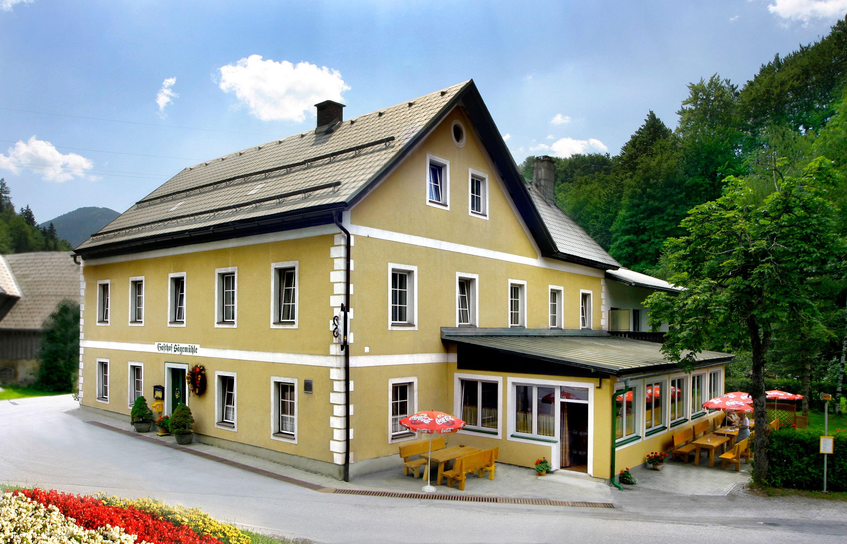 A yellow inn with a terrace and parasols, surrounded by trees.
