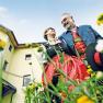 A couple in traditional costume stand in front of a yellow building surrounded by flowers, smiling at each other.