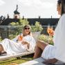 Two women in bathrobes relax on deckchairs with drinks in front of a castle.