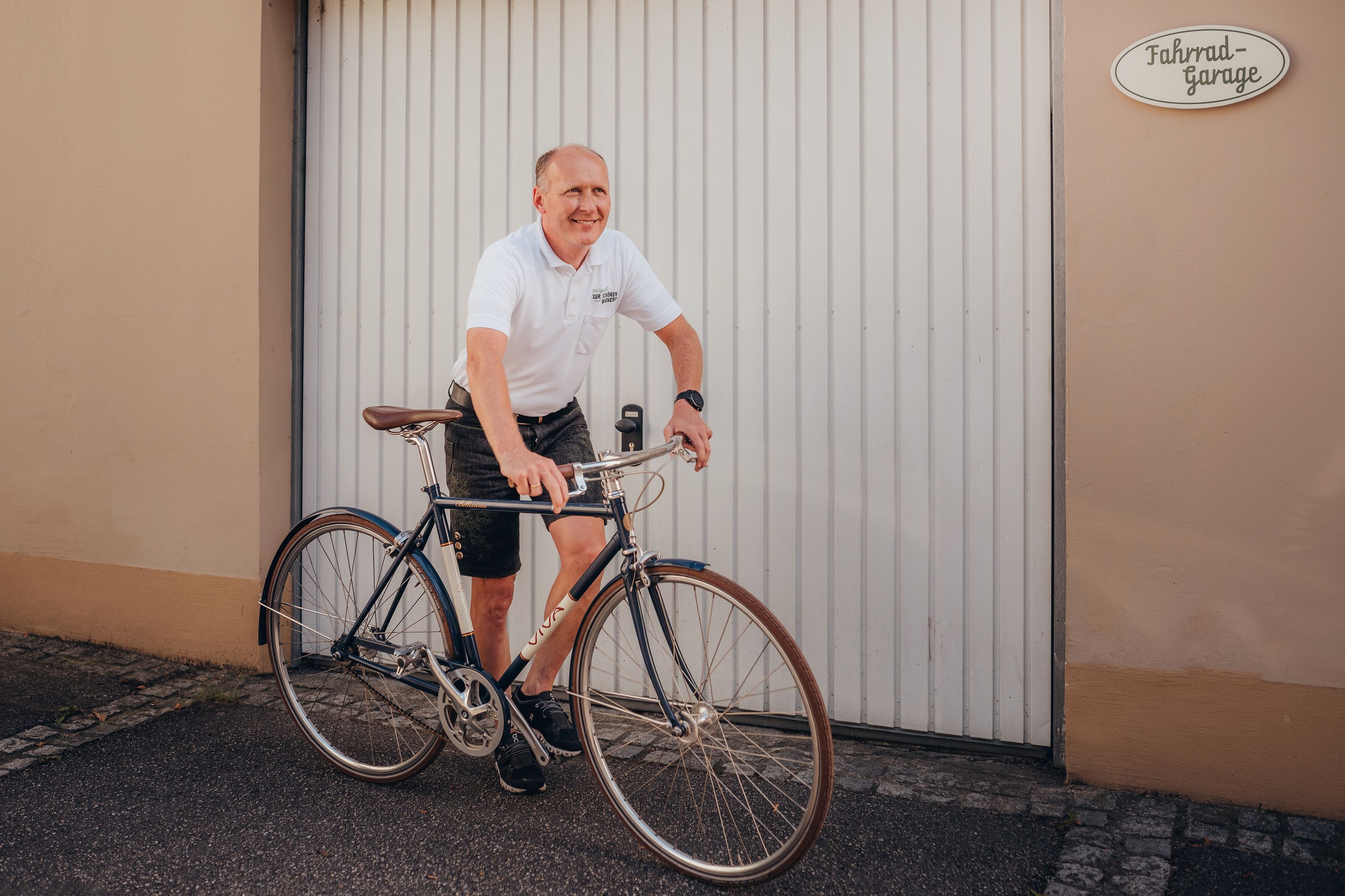 Man in white polo shirt riding bicycle in front of garage door.