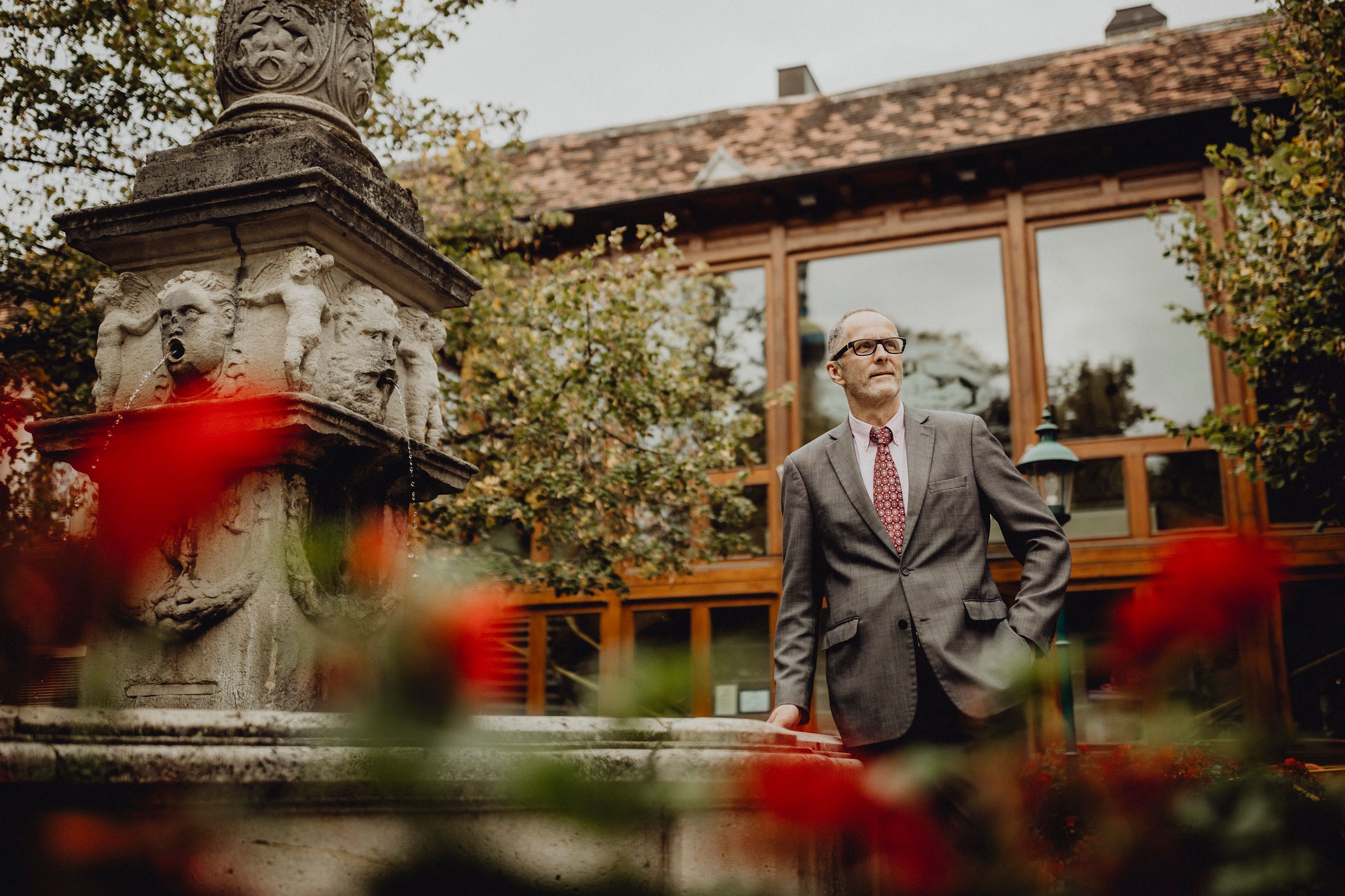 A man in a suit stands next to a stone fountain with gargoyles, surrounded by red flowers and a building in the background.