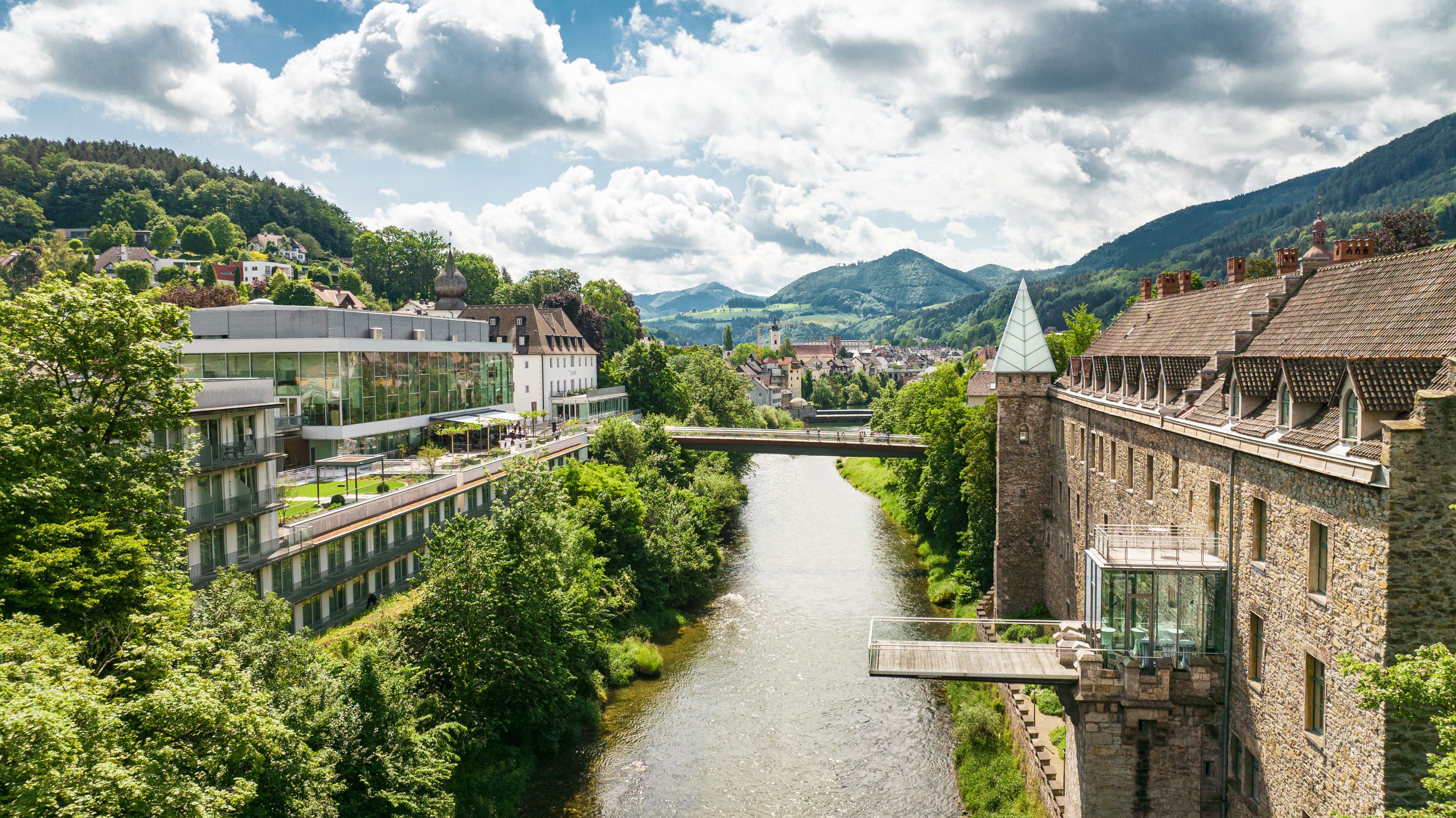 Panorama of a river landscape with a castle and modern architecture, surrounded by green hills.