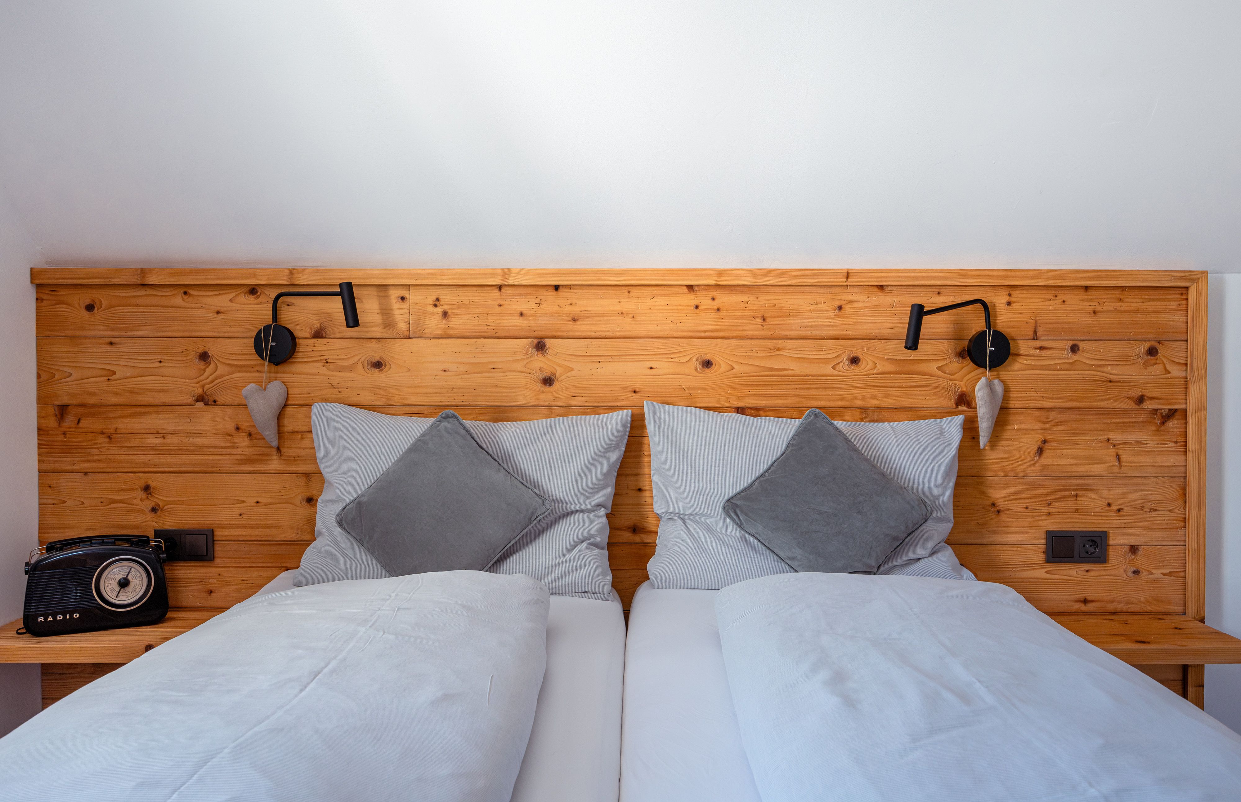 Comfortable double bed with wooden back panel, gray cushions and blankets. A retro radio on the left, a reading lamp and heart decoration on the right.