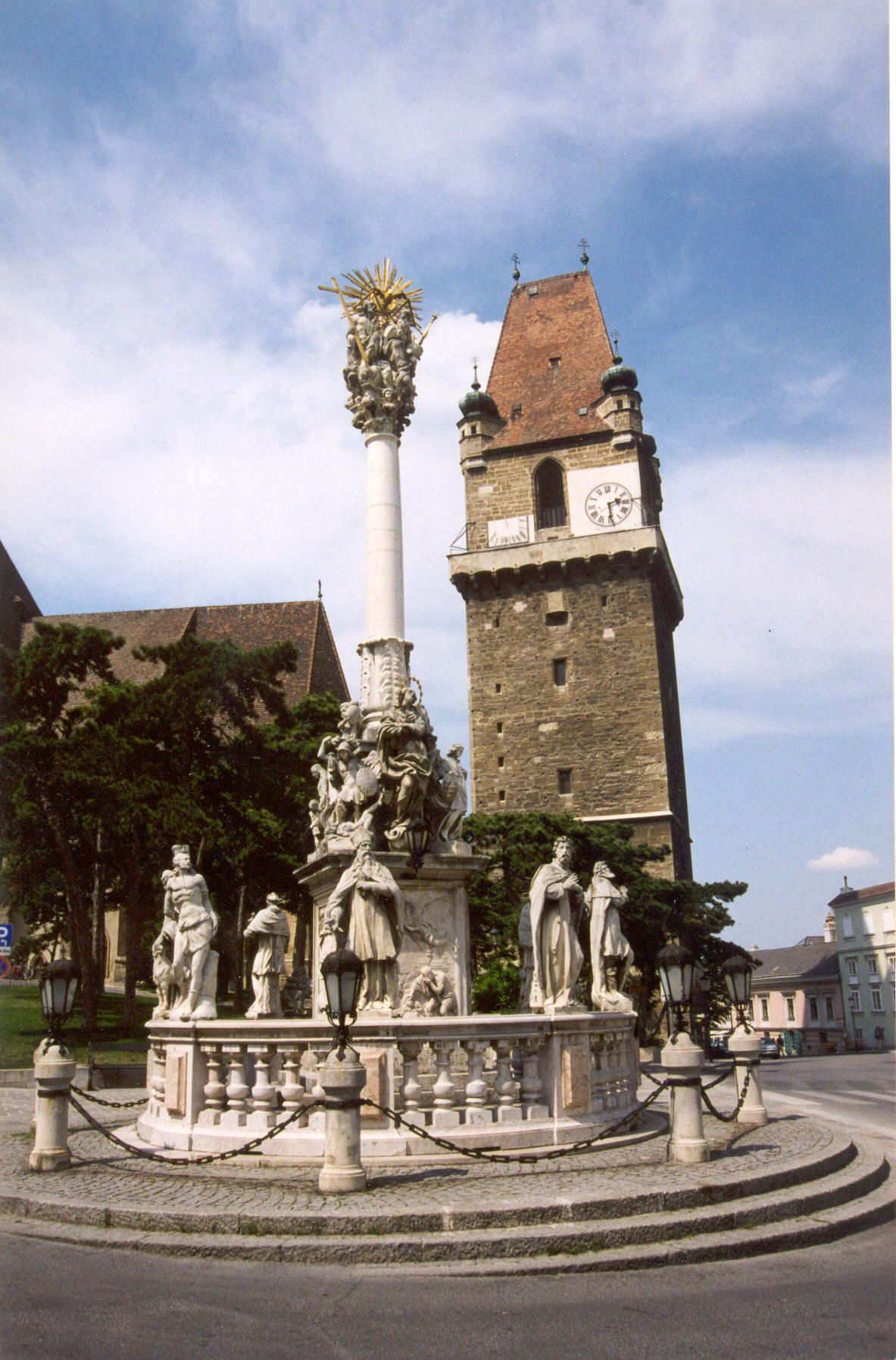 Historic defense tower with clock and baroque column in the foreground.