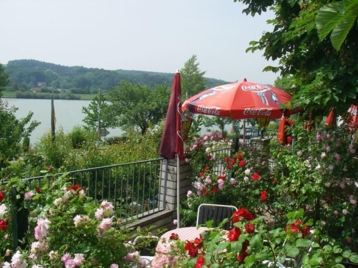 A blooming garden with red and pink roses, a table with chairs and a red parasol with the Coca-Cola logo, with a lake in the background.