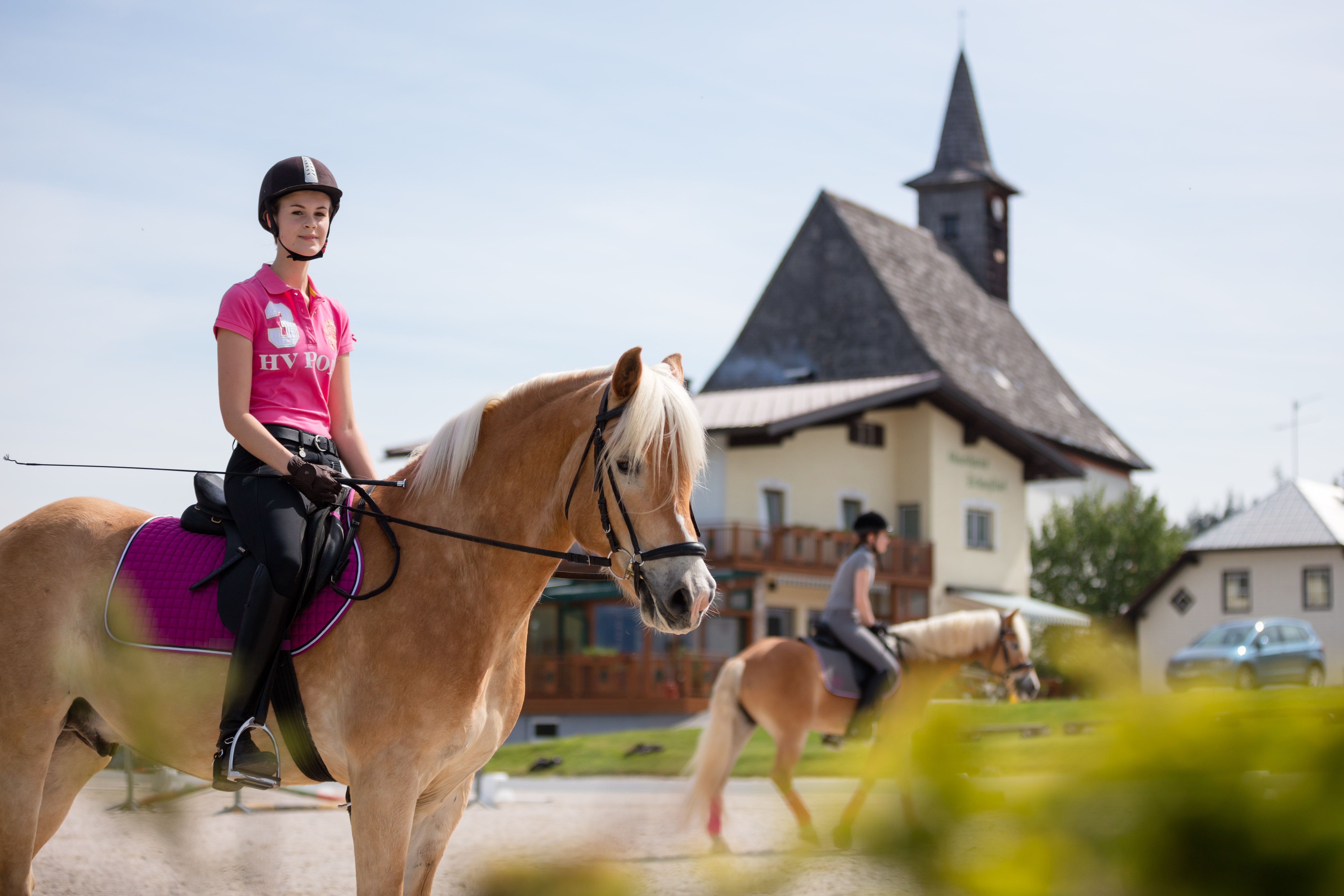 Two female riders on horses in front of a building with a tower.