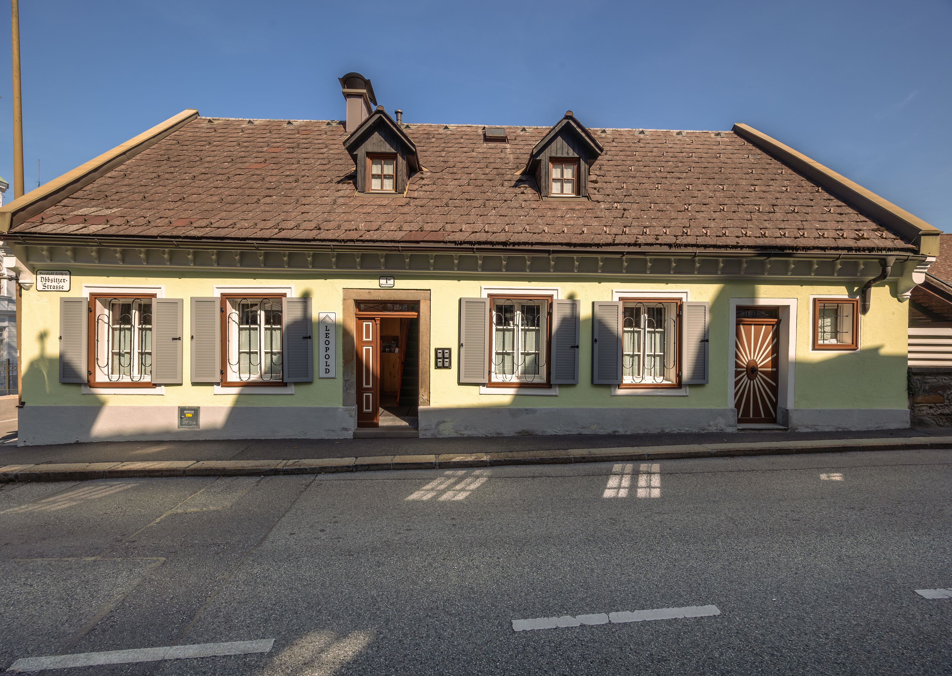 A traditional yellow house with a brown roof and green shutters on a street.