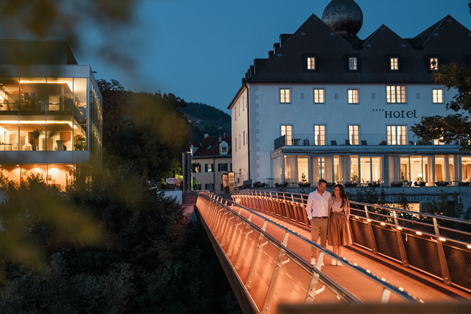 A couple walks across an illuminated bridge in front of a hotel in the evening.
