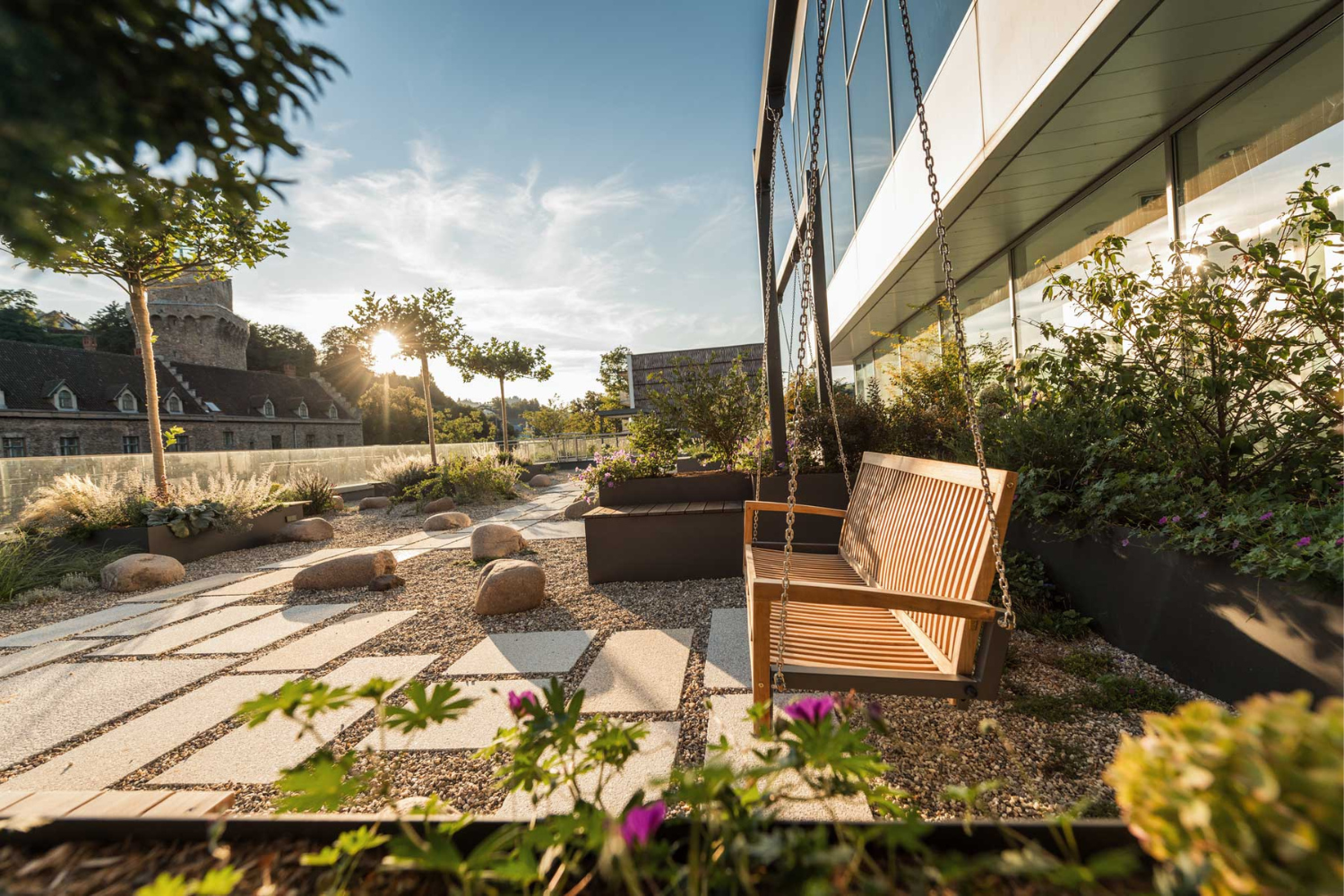 Garden with rocking bench, gravel path and plants, in the background a castle at sunset.