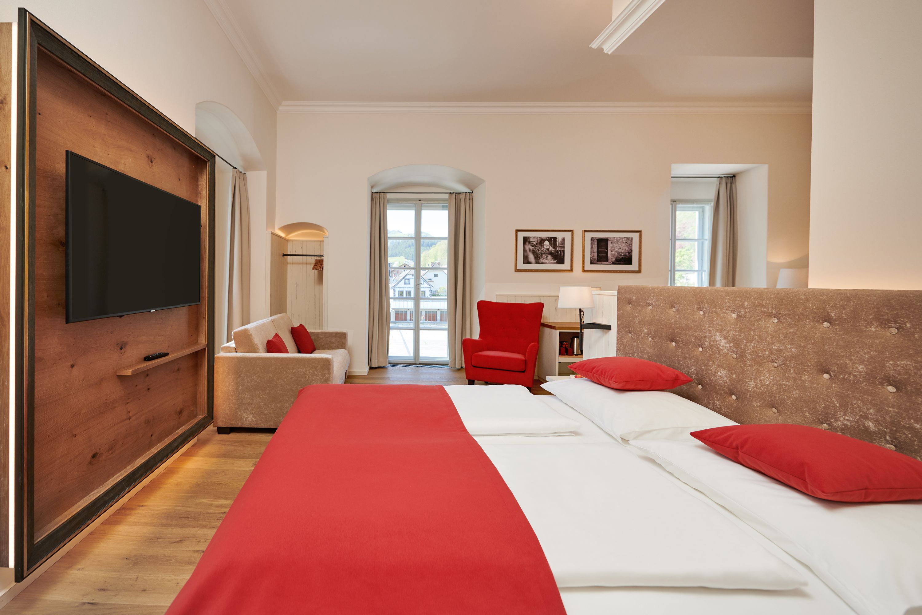 Hotel room with double bed, red bed linen, sofa, red armchair and large window.
