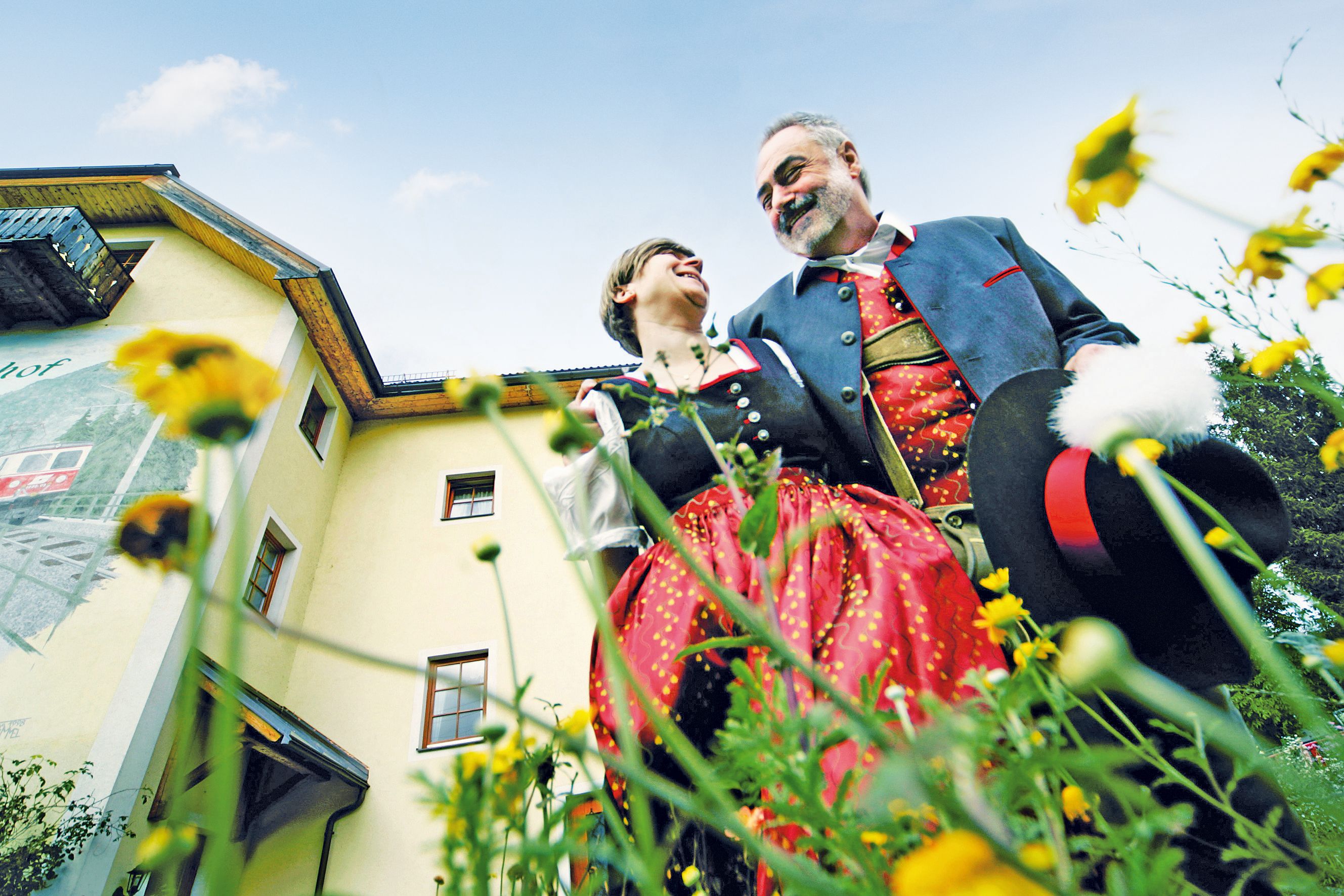 A couple in traditional costume stand in front of a yellow building surrounded by flowers, smiling at each other.