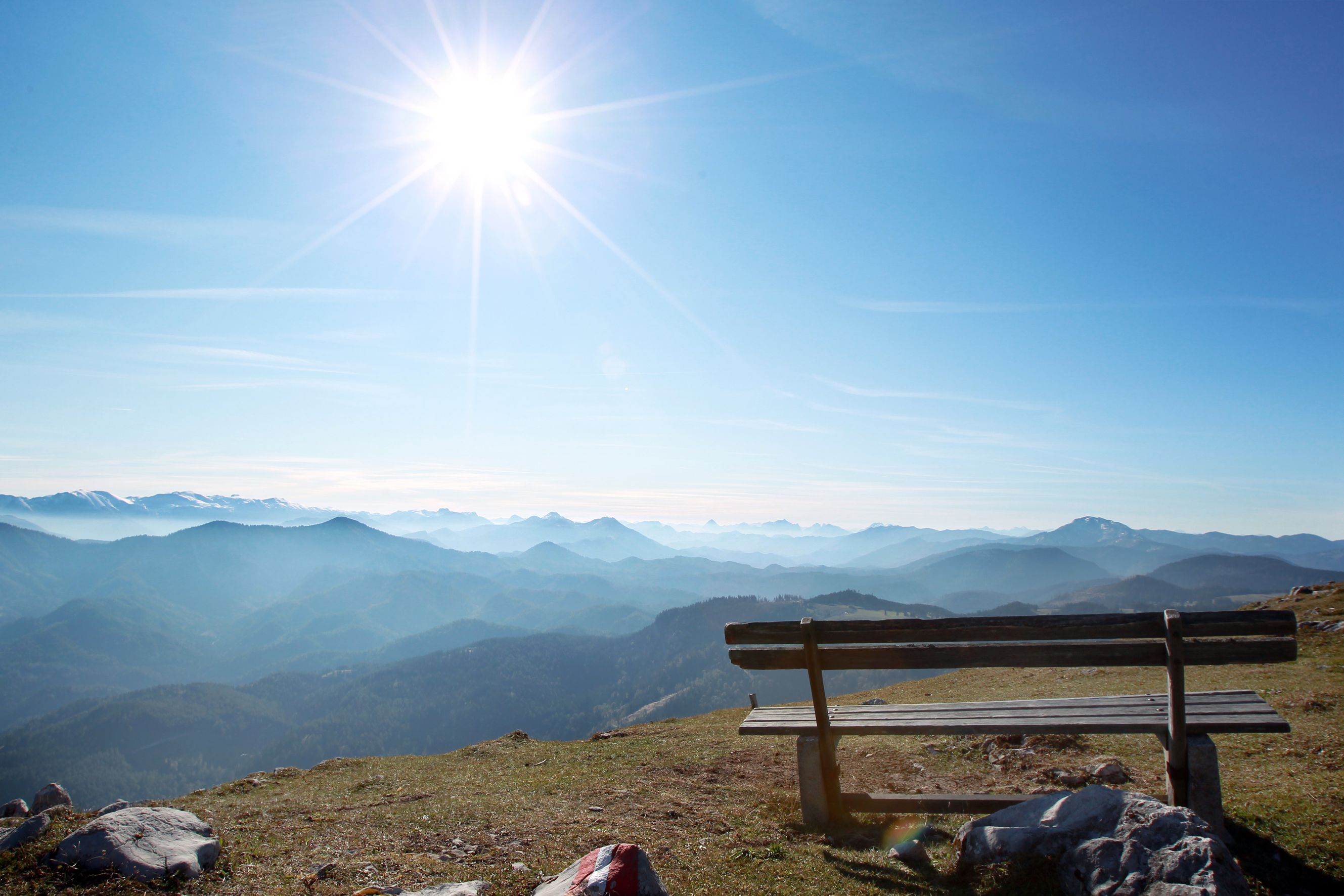 Wooden bench on a mountain with a view of a mountain landscape and the sun in the sky.