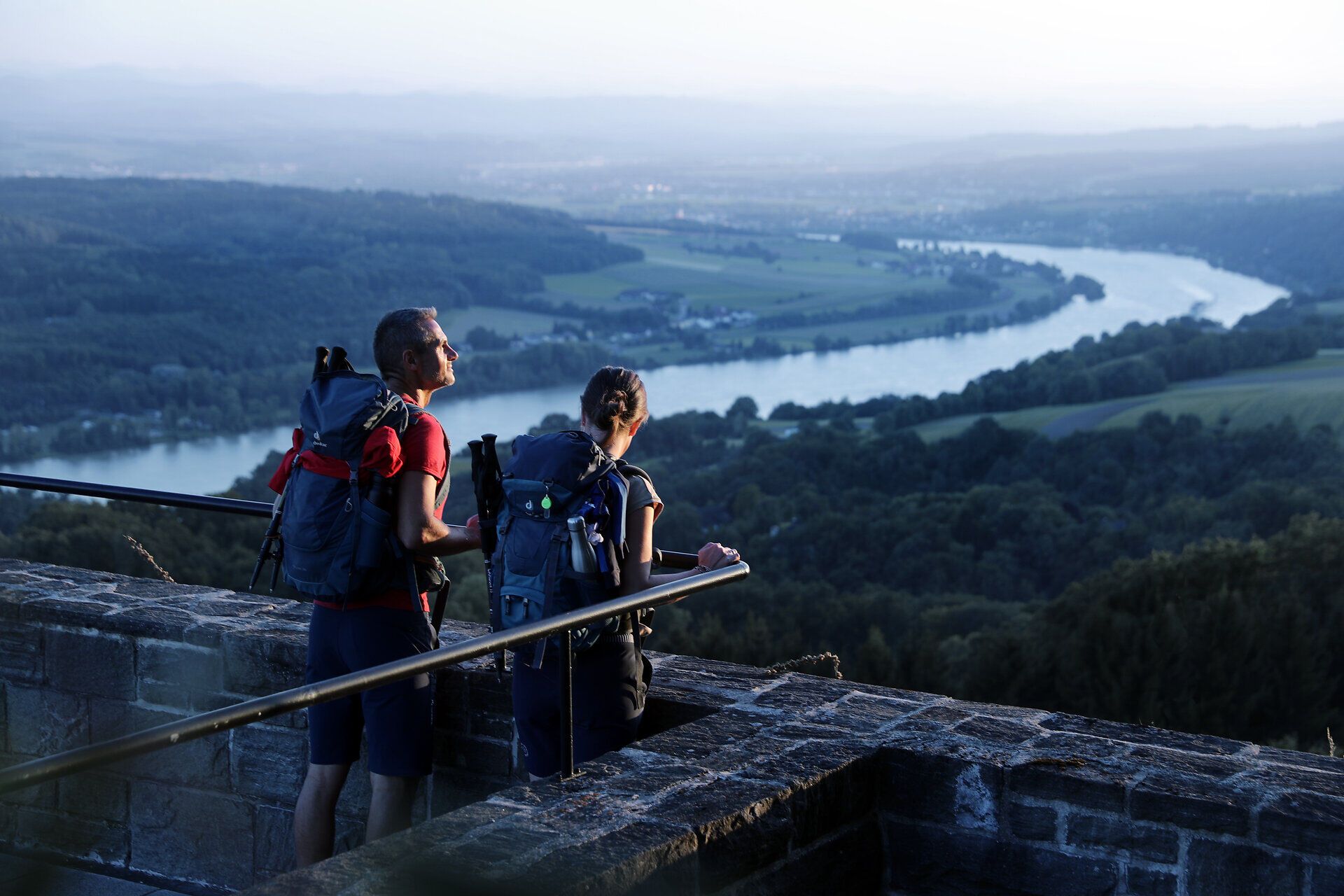 Die sanften Hügel des Mostviertels erstrecken sich majestätisch im Hintergrund, während zwei Wanderer die atemberaubende Aussicht genießen. Die ruhige Atmosphäre und die frische Bergluft laden dazu ein, die Seele baumeln zu lassen und die Schönheit der Natur zu erleben.