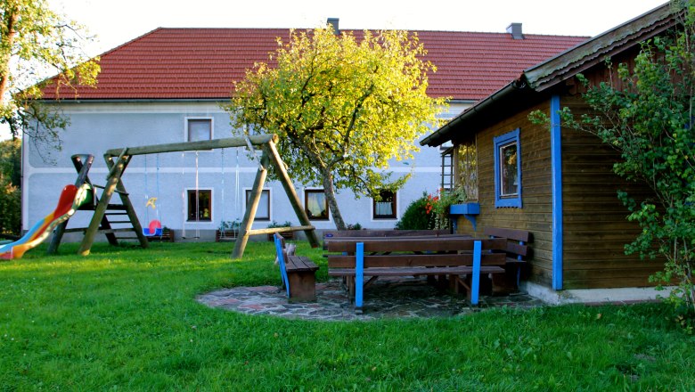 A playground with a swing, slide and benches in front of a house with a red roof.