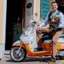 A man sits on a scooter in front of a building holding a tray with beer bottles and a glass.