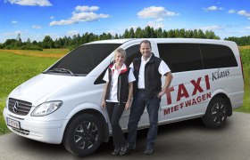 Two people are standing in front of a white Mercedes van labeled 'Taxi Mietwagen Klaus' on a rural road.