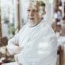 A man in chef's clothes stands with his arms folded behind a pane of glass.