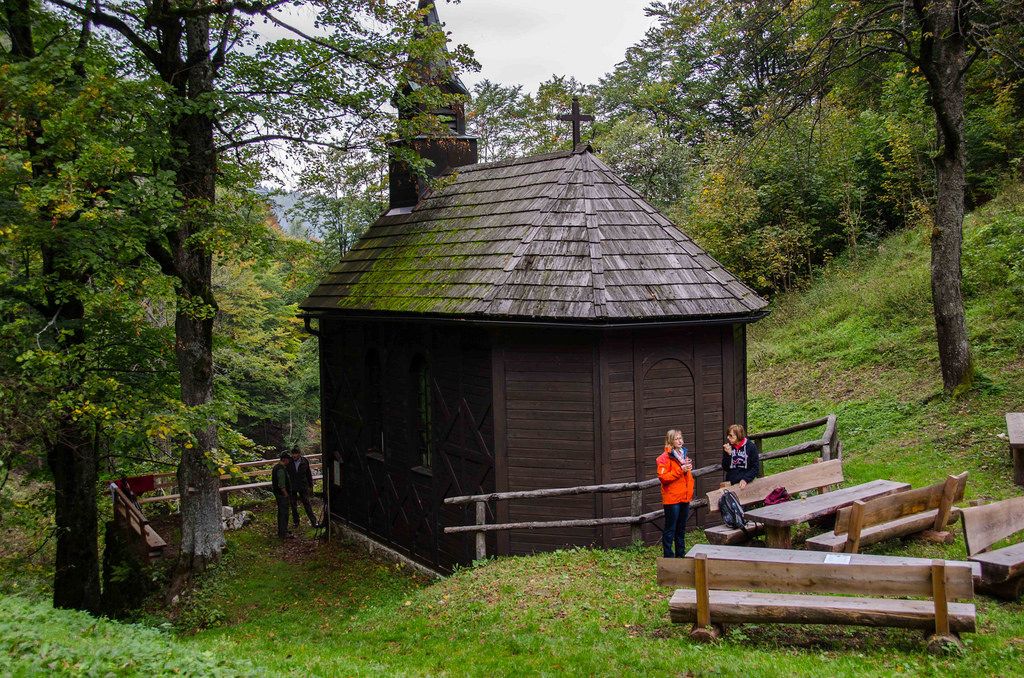 A small, wooden church in the forest with two people and wooden benches in the foreground.