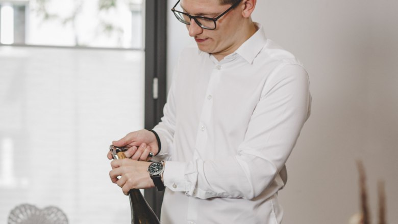 A man opens a bottle of wine at a table set with food.