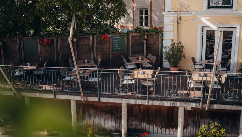 Cozy terrace of a caf&eacute; with wooden tables and chairs, surrounded by plants.