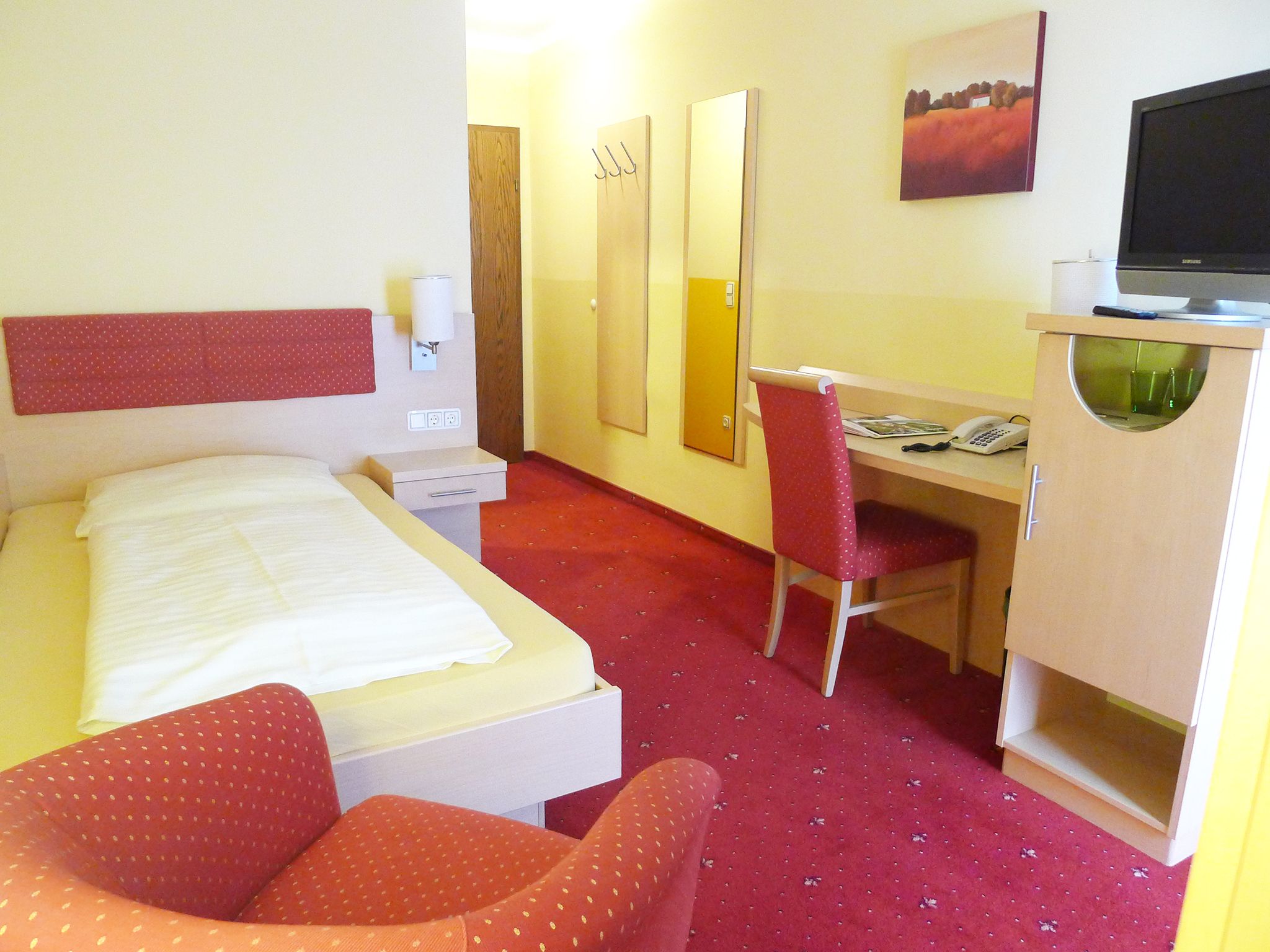 A hotel room with a single bed, red carpet, desk and chair. A picture hangs on the wall, next to it a television on a cupboard.