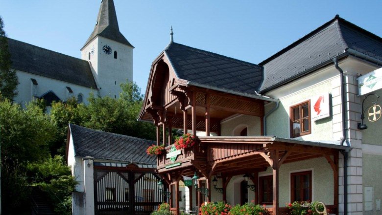 A traditional building with a wooden balcony and floral decorations, next to a church with a tower.